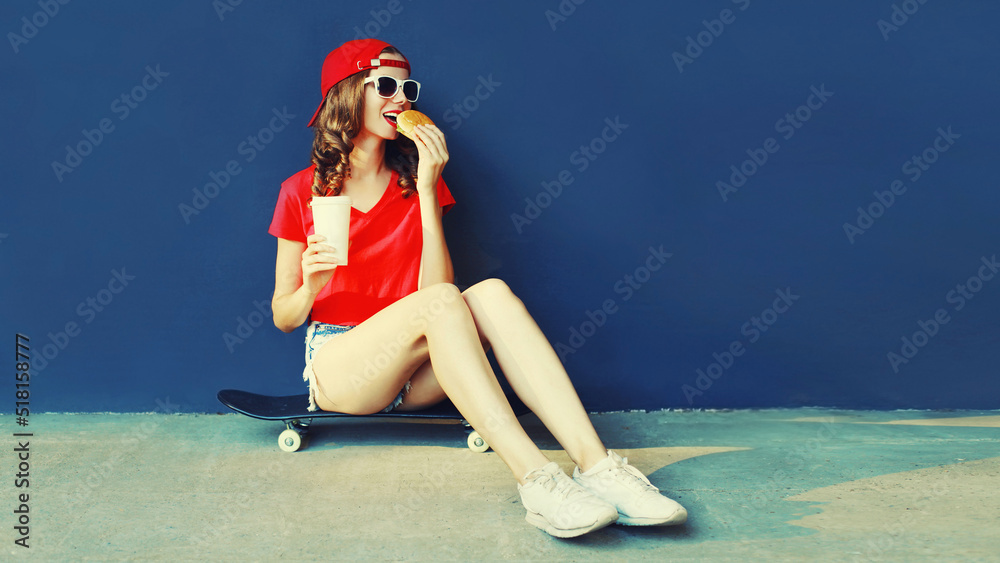 Wall mural cool young woman with burger and coffee cup sitting on skateboard wearing baseball cap, sunglasses o