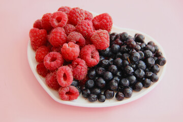 ripe raspberries and blueberries on a white plate in the shape of a heart on a light background close-up