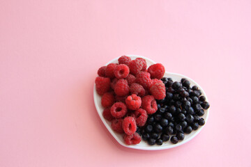 ripe raspberries and blueberries on a white plate in the shape of a heart on a pink background