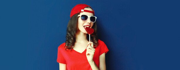 Portrait of young woman with heart shaped red lollipop wearing baseball cap, sunglasses on blue...