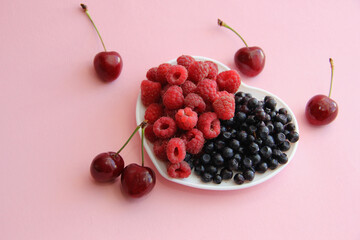 Large raspberries and blueberries on a white plate in the form of a heart on a pink background