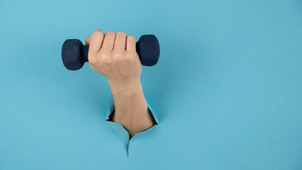 A female hand sticking out of a hole from a blue background holds a dumbbell. 