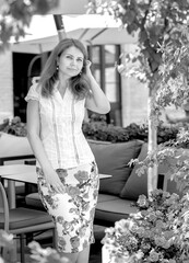 Black and white portrait brunette girl in a white blouse and midi skirt with a rose print standing in a summer cafe, playground with flowers. Looking into the camera with a smile. Summer concept