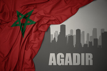 abstract silhouette of the city with text Agadir near waving colorful national flag of morocco on a gray background.