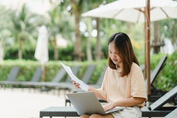 Woman working by the pool with a laptop computer holding documents in hand. Asian women's holiday...