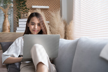Young Asian woman using laptop on sofa living room at home cozy happy working at home.