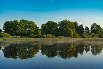 Fototapeta na wymiar Landscape view on the river and coast with trees in morning light and reflection in water