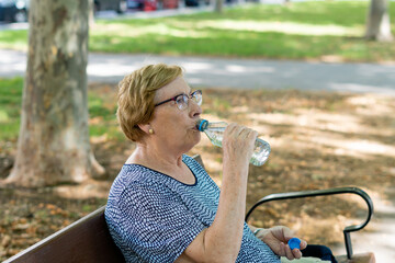 Portrait caucasian elderly woman drinking water from a plastic bottle, sitting on a park bench.