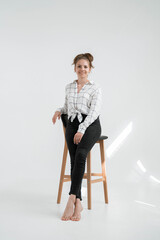 Fototapeta premium A sexy slender girl with hair in a bun is stylishly dressed in a shirt and pants, isolated on grey background. A woman sits on a high chair in a photo studio