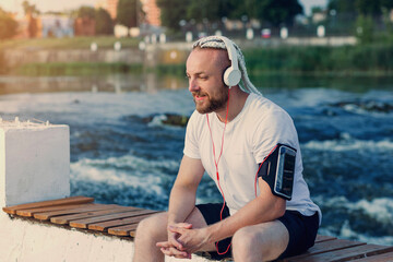 A man with a beard and braid hair is listening to music with headphones on the river bank and squinting from the sun