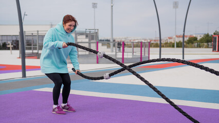 Fototapeta na wymiar Caucasian woman in a mint sweatshirt is training with battle ropes at the sports ground. 