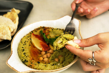 Hummus with avocado in a ceramic pan and Lebanese tortilla. Traditional Jewish cuisine. A woman's hand dips piece of bread into hummus.