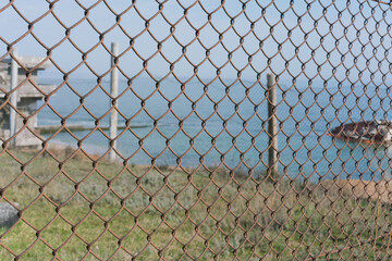 A metal rusty grate fence near a sea coastline. Wire fence. Fencing. Guard. Defense. Safeguard. Shield. Netting. Material. Manufacture. Limit. Restriction. Stop. Control. Regulation. Jail