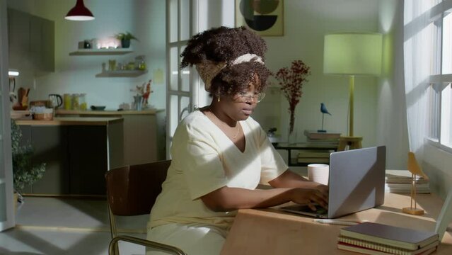 Zoom in shot of young African American woman in glasses working on laptop at desk in evening at home
