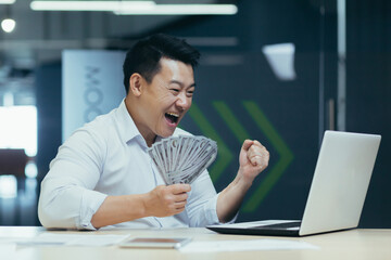 Successful Asian investor businessman, happy with victory and conclusion of investment contract, man holds dollars cash in hand and shouts joyfully at laptop screen