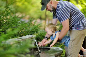 Father helping his toddler son to wash his hand in city fountain.
