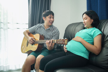 Fototapeta na wymiar Asian husband and pregnant wife spend time together in the bedroom and husband play guitar sing-song for an unborn baby at home.