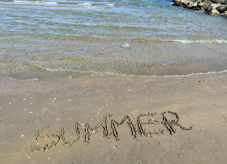 word SUMMER in capital letters on the sand of the sandy beach by trhe sea