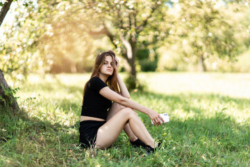 Naklejka na ściany i meble a young girl in a black shirt and black shorts sits on the grass in the park in summer holding her phone and looking at the camera. photo with sun glare