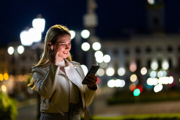 Smiling young woman walking in the city at night with headphones enjoy listening online music from smartphone and looking illuminated city night lights. People and technology concept.