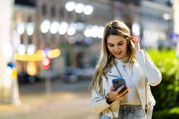 Beautiful young stylish woman walking in the night city streets using modern smartpone