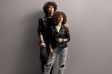 Young man and woman with leather jackets and curly hair leaning on a gray wall
