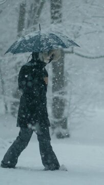 SLOW MOTION, DOF: Young woman wearing a thick winter coat and carrying umbrella walks through a blizzard during a relaxing stroll in the picturesque wintry park. Female walks into fresh powder snow.