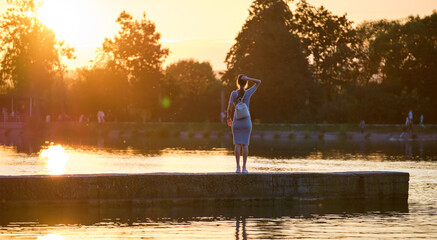 Back view of lonely young woman standing alone on lake shore enjoying warm evening. Wellbeing and relaxing in nature concept