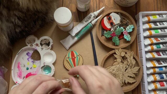 Fluffy cat on wooden table watches women's hands mix acrylic paint with gold sequins in palette. Tubes of color, brush, brooches made of plywood, badges in form of branches, Santa, aerostat and pizza.
