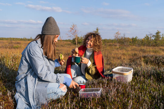 Two cheerful young girls having picnic outdoors during sunny autumn day. Happy girlfriends spend fall weekend in countryside eating sandwiches and drinking hot tea or coffee with happy relaxed smiles