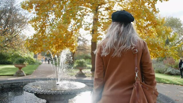 blonde English lady with dark blue hat standing by a fountain in the botanical garden in Oxford Uk, then turning around and smiling into the camera