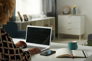 Close-up of African girl working online using her laptop while sitting at table with notepad and cup of tea in living room