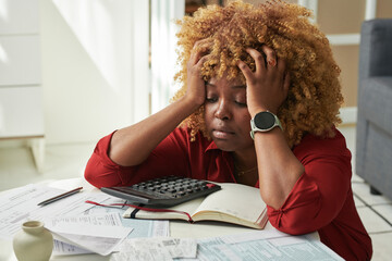 African exhausted woman holding her head while sitting at table with bills and taxes, she tired of...