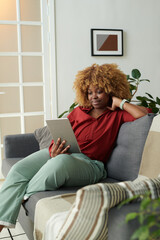 African girl resting on sofa in living room and having online communication on digital tablet