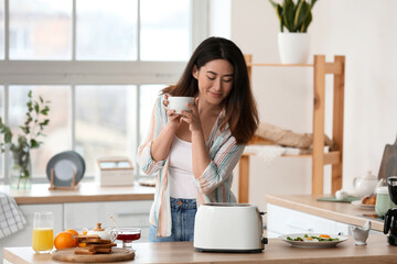 Beautiful young Asian woman drinking coffee and making tasty toasts in kitchen