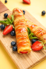 Board with thin pancakes, berries, jam and mint leaves on yellow background, closeup