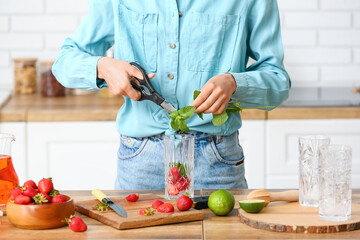 Woman adding fresh mint into glass with strawberry lemonade at table in kitchen