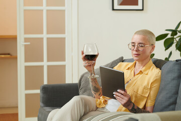 Young woman relaxing on sofa with glass of red wine and enjoying of watching video on digital tablet