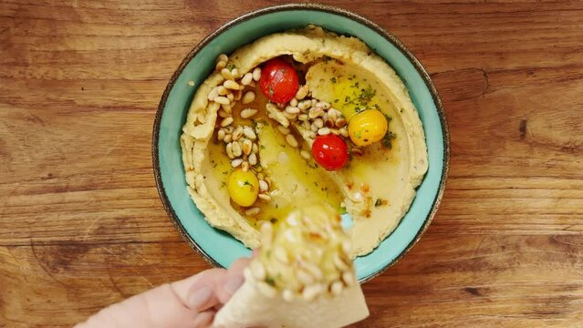A woman's hand scoops up a piece of bread Delicious hummus with cherry tomatoes and pine nuts with a tortilla of pita bread, which lies in a plate of sea-green, standing on a wooden table. Top view.