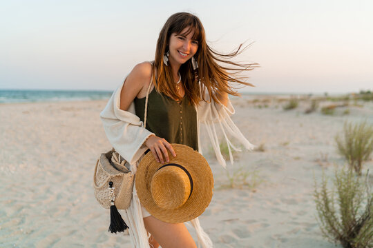 Summer photo of happy young woman in stylish boho  outfit  holding straw bag and hat  posing on tropical beach. Windy hairs.