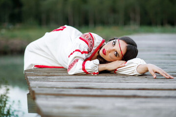 A Slavic girl is lying on a wooden pier in national Ukrainian clothes .