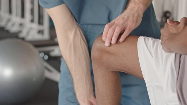 Midsection of unrecognizable physiotherapist in blue scrubs observing shin bone of Black male patient having knee injury during physiotherapy session