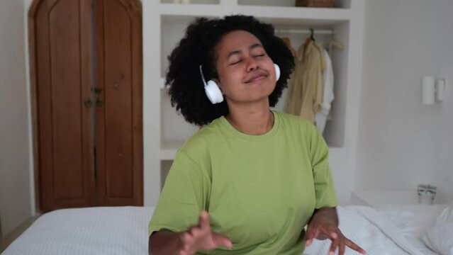 A mixed-race woman with an Afra hairstyle moves to the beat to the incendiary music from her headphones. Afro-Asian woman with obsession listens to music in the bedroom on the bed.