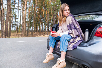 Young woman sitting on car trunk with coffee cups and thermos on the side of the road in the forest in warm fall or spring day. Time for yourself. Contemplation and rest in solitude.