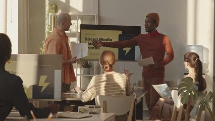 Medium slowmo of two young creative diverse men making presentation on SMM trends to group of colleagues, working on product together in successful digital marketing agency