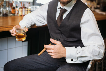A unrecognizable  man in an elegant vest holds a glass of whiskey near bar counter. Toast at the...