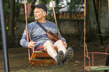 Carefree senior man in casual clothes and black cap having fun and swinging on swing in amusement...