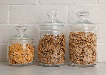 Glass containers with different breakfast cereals on white countertop near brick wall