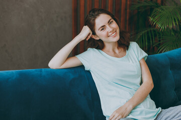 Young happy smiling cheerful calm fun european woman she 20s wearing casual clothes look camera prop up head relax sit on blue sofa indoor rest at home in own room apartment. People lifestyle concept.