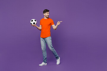 Full size young fan man he wear orange t-shirt cheer up support football sport team hold soccer ball watch tv live stream point index finger aside on workspace isolated on plain dark purple background